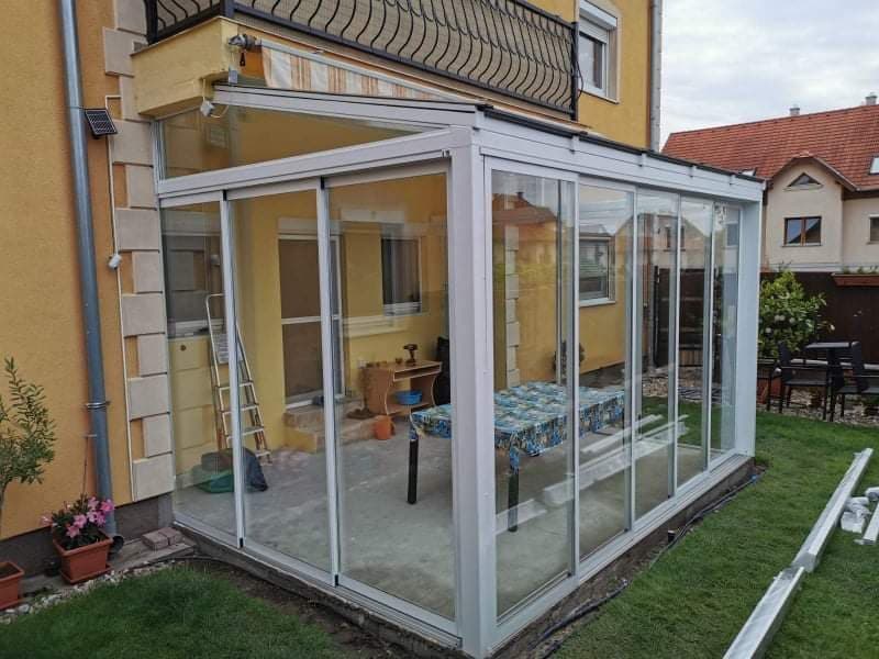 Fehér alumínium vázas, üvegfalú terasz egy sárga családi ház udvarán, tolható üvegoldalakkal.
