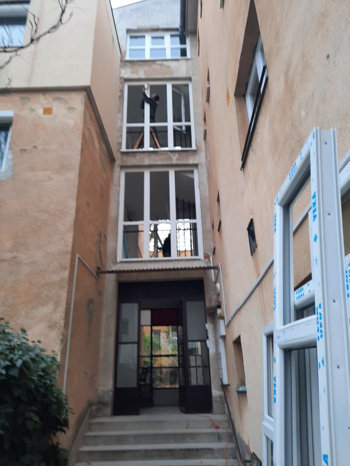 Lépcsőházi ablakcsere az udvar felől, a nyílásban új fehér ablakszerkezet, mellette a következő tok fóliában.